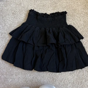 Svart volangkjol med smock - Supersöt svart kortkjol med bred smock i midjan och dubbla volanger. Kjolen har en luftig känsla och passar perfekt till sneakers eller boots. Enkel att styla med crop top eller stickad tröja. Storlek 140🥰