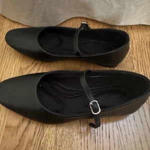 Svarta ballerinaskor med rem - Stilrena svarta ballerinaskor med smal rem och silverfärgat spänne över foten. Skorna har rund tå och platt sula, tillverkade i ett slätt syntetmaterial som ger en klassisk look. Perfekta för dig som gillar enkel och tidlös stil.