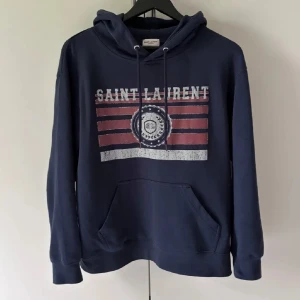 Saint Laurent marinblå hoodie med tryck - Snygg marinblå hoodie från Saint Laurent med vit och röd vintageinspirerad logga och ränder på bröstet. Tröjan har huva med snörning, känguruficka och långa ärmar. Perfekt för dig som vill ha en streetig och exklusiv look.(Storlek M men passar mer som S)