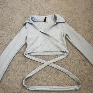 Ljusblå knytbar topp - Supersnygg ljusblå omlottopp från Divided i ribbad bomullsmix. Toppen har långa ärmar, croppad passform och långa knytband som kan snurras runt midjan. Perfekt för dig som gillar trendiga detaljer och stilren look.