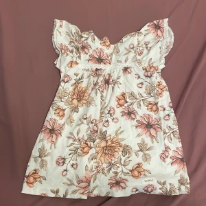 Blommig kortklänning från Newbie - Supersöt kortklänning från Newbie med romantiskt blommönster i beige, rosa och bruna toner. Klänningen har volangärmar, spetsdetaljer och knappar i ryggen. Perfekt för varma dagar och somriga tillfällen.