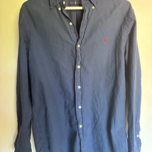 Blå skjorta från Ralph Lauren M - Säljer en klassisk blå skjorta från Ralph Lauren i storlek M. Skjortan har långa ärmar, vita knappar och den ikoniska röda loggan broderad på bröstet. Tillverkad i mjuk bomull och passar perfekt till jeans eller shorts.