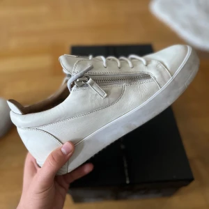 Vita sneakers från Giuseppe Zanotti - Snygga vita sneakers från Giuseppe Zanotti i skinn med cool blixtdetalj på plösen och dragkedja på sidan. Klassisk rund tå, snörning och platt sula. Perfekta för dig som gillar stilrena och exklusiva sneakers med unika detaljer.  Jag köpte dem för 3,5k sek nya. Skolådan kommer med med alla kort och brev som de skickade. Använda ett 5 gånger kanske men eftersom dem är vita ser de lite använda ut