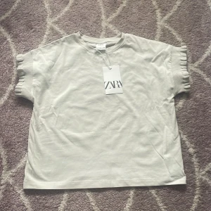 Vit topp med volangärm från Zara - Superfin vit topp från Zara med korta ärmar och snygga volangdetaljer vid ärmsluten. Enkel och stilren design som passar perfekt till jeans eller kjol. Tillverkad i mjuk bomull för en skön känsla hela dagen.Och helt oanvänd och är i storlek 140.