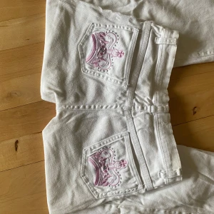 Vita jeans med rosa kronbrodyr - Unika vita jeans från Stretch med snygg rosa kronbrodyr på bakfickorna och en mindre krona vid fickan fram. Jeansen har klassisk femficksmodell, raka ben och silverfärgade detaljer. Perfekta för dig som vill sticka ut med en cool detalj.
