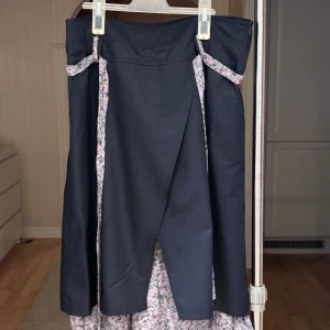 Svart omlottkjol med blommigt foder - Unik svart långkjol med omlott-design och blommigt foder i grått, rosa och grönt. Kjolen har dekorativa knytband i samma blommiga tyg och volangkant nertill. Perfekt för dig som gillar kontraster och romantiska detaljer.