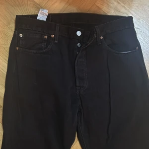 Svarta Levi's 501 jeans W34 L32 - Klassiska svarta Levi's 501 jeans med raka ben och knappgylf. Jeansen har fem fickor, silverfärgade knappar och den ikoniska läderpatchen bak. Tillverkade i slitstarkt bomullstyg, perfekt för en clean och tidlös look.