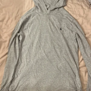 Grå hoodie från Polo Ralph Lauren 16-18 år  - Snygg ljusgrå hoodie från Polo Ralph Lauren i storlek XL. Tröjan har lång ärm, huva och klassisk broderad logga på bröstet. Tillverkad i mjuk bomullsmix som är skön att ha på sig. Perfekt för en chill och stilren look. Helt oanvänd. Storlek XL i barn (170-180) passar Xs & S