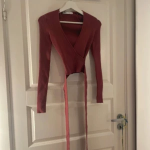 Ribbad omlottop från Zara i röd - Supersnygg ribbad omlottop från Zara i en mörkröd nyans. Långärmad med djup v-ringning och långa knytband i midjan. Stretchigt material som sitter tajt och framhäver figuren. Perfekt till jeans eller kjol för en trendig look.
