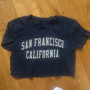Mörkblå croppad t-shirt J.Galt - Mörkblå croppad t-shirt från J.Galt med vit text 'San Francisco California' på bröstet. Klassisk rund halsringning och korta ärmar. Skön bomullskvalitet och avslappnad passform, perfekt till jeans eller shorts.