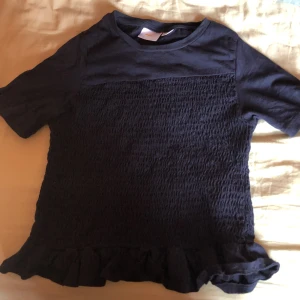 Mörkblå topp med volang från Gina Tricot - Supersöt mörkblå topp från Gina Tricot med korta ärmar och smockad front. Toppen har en volangkant längst ner som ger en extra touch. Perfekt till jeans eller kjol för en chill look.