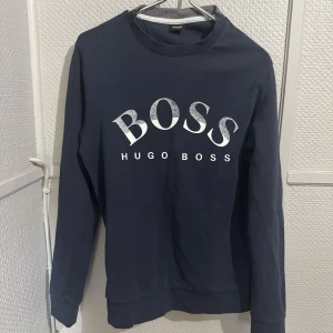 Mörkblå sweatshirt från Hugo Boss - Snygg mörkblå sweatshirt från Hugo Boss med stor vit BOSS-logga framtill. Tröjan har rund halsringning och långa ärmar. Tillverkad i mjuk bomull som är skön att ha på sig. Perfekt för en clean och stilren look.