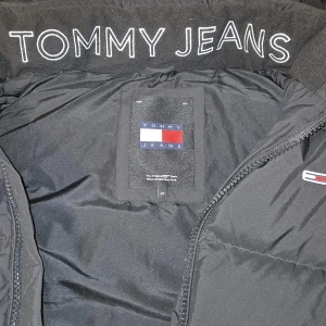 Svart dunväst från Tommy Jeans - Säljer en svart dunväst från Tommy Jeans med broderad logga på bröstet och stor TOMMY JEANS-text på insidan av kragen. Västen har huva och dragkedja framtill, perfekt för lager-på-lager-stil. Klassisk och stilren design som funkar till det mesta.