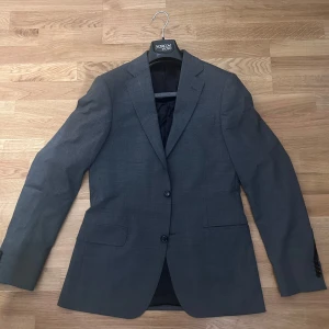 Mörkgrå kavaj från Oscar Jacobson - Snygg mörkgrå kavaj från Oscar Jacobson med klassisk passform och två knappar framtill. Kavajen har innerfoder i svart, flera innerfickor och raka slag. Köpt Maj 2025. Nyskick, utan lappar. 