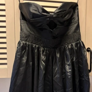 Svart axelbandslös kort klänning med rosett - Supersöt svart axelbandslös kort klänning i satin med stor rosettdetalj framtill och cut-out under bysten. 