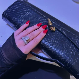 Svart kuvert/clutch väska i skinn med struktur - Säljer en svart kuvertväska i skinn med snygg ormskinnsstruktur och guldfärgad vridknäppning. Väskan har stilren design och ett smalt handtag, perfekt för att lyfta din outfit med en edgy touch. Endast använt en gång. Fick många komplimanger