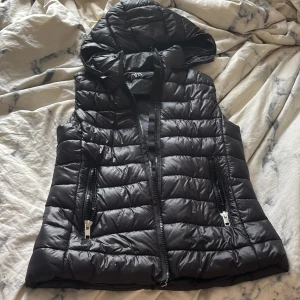 Svart dunväst med huva från Zara - Svart quiltad dunväst från Zara med huva och två fickor med dragkedja. Västen har en rak passform och blanka detaljer. Perfekt att slänga över en hoodie för en cool look.