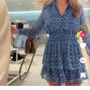 Blå mönstrad kortklänning med volang - Supersöt blå kortklänning med vitt mönster och volangkant. Klänningen har långärmade transparenta ärmar, markerad midja med smock och v-ringad krage. Perfekt för dig som gillar flowiga och luftiga plagg med snygg passform.