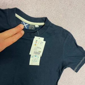 Mörkblå t-shirt från Lindex - En basic mörkblå t-shirt från Lindex med rund halsringning och korta ärmar. Tillverkad i mjuk bomull och har diskreta kontrastsömmar vid halsen. Perfekt för en clean och enkel look.