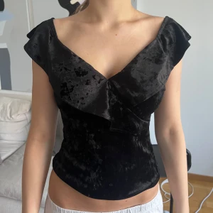 Svart sammetstopp med volang - Supersnygg svart topp i mjuk sammet med v-ringning och volangdetalj framtill. Toppen är ärmlös med breda axelband och har en figurnära passform som ger en elegant siluett. Perfekt för dig som vill sticka ut med en lyxig känsla.