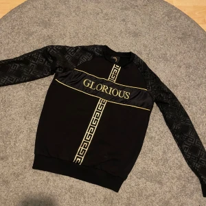 Svart tröja med guldtryck GLORIOUS - Svart långärmad tröja från Glorious med guldiga detaljer och tryck. Snyggt mönster på ärmarna och guldiga linjer samt texten GLORIOUS framtill. Tröjan har ribbade muddar och ett exklusivt streetwear-inspirerat utseende.