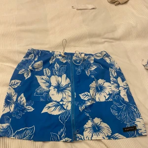 Blå blommig badkjol från Esprit - Snygg blå badkjol  från Esprit med stora vita hibiskusblommor. Kjolen har snörning i midjan, dragkedja framtill och en bakficka. Perfekta för stranden eller poolen, med en riktigt somrig vibe och skön passform.