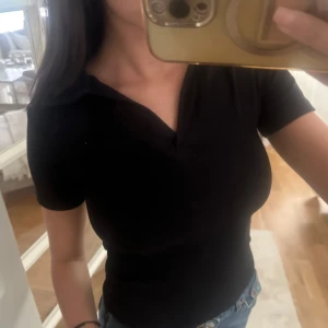 Svart ribbad polotopp från Refresh - Svart ribbad polotopp från Refresh med korta ärmar och snygg krage med v-ringad öppning. Toppen har en tight passform och är gjord i stretchigt material som sitter skönt på kroppen. Perfekt till jeans eller kjol för en clean look. Storlek S