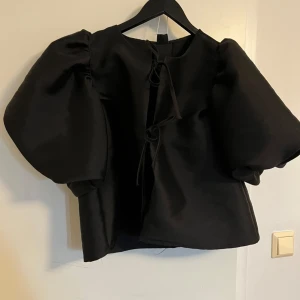 Svart blus med puffärm från Gina Tricot - Supersnygg svart blus från Gina Tricot med stora puffärmar och trendiga knytningar framtill. Blusen har en boxig passform och är perfekt för dig som vill sticka ut med en cool och modern look. Materialet är slätt och ger en lyxig känsla.
