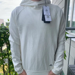 Vit hoodie från C.P. Company - Snygg vit hoodie från C.P. Company med deras ikoniska goggles på huvan. Tröjan har en clean look, lång ärm och dragkedja i sidan. Perfekt för dig som vill sticka ut lite extra. Tillverkad i mjuk bomull för skön känsla. Storlek  xs, men passar dig som är från 165-175