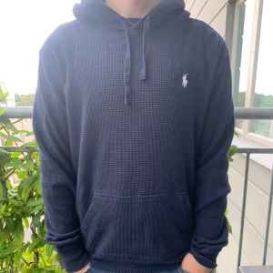 Mörkblå hoodie från Polo Ralph Lauren - Säljer en mörkblå hoodie från Polo Ralph Lauren med vit broderad logga på bröstet. Tröjan har huva med snörning, känguruficka och är tillverkad i ett mjukt, stickat bomullstyg med diskret rutstruktur. Perfekt för chill dagar eller när du vill vara extra bekväm.