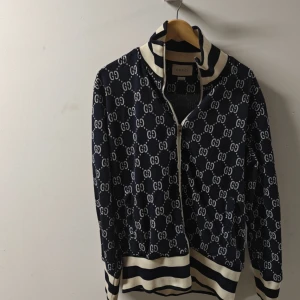 Gucci trackjacka med GG-mönster xl - Säljer en blå och vit trackjacka från Gucci med klassiskt GG-mönster över hela jackan. Jackan har dragkedja framtill, vita och röda ränder längs ärmarna samt ribbade muddar i vitt och blått. Perfekt för dig som vill sticka ut med en ikonisk streetstyle-look.