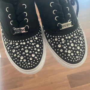Svarta sneakers med nitar från Guess - Svarta sneakers från Guess med vita sulor och snörning. Skorna är dekorerade med massor av silvriga nitar och stenar över hela ovandelen för en riktigt blingig look. Guess-logga på plösen och hälen. Perfekta för dig som vill sticka ut.