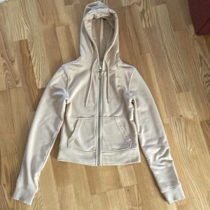 Beige croppad hoodie från H&M Divided - Säljer en beige croppad hoodie från H&M Divided i storlek XS. Tröjan har dragkedja framtill, huva med snörning och två fickor. Perfekt för en chill och avslappnad stil. Tillverkad i mjuk bomullsmix som är skön att ha på sig.