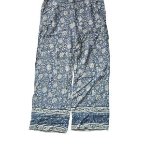 Blommiga blå byxor från H&M - Säljer ett par blå byxor från H&M med vitt blommönster och små röda detaljer. Byxorna har avslappnad passform, resår i midjan och är tillverkade i mjuk bomull. Perfekta för en chill och stilren look.