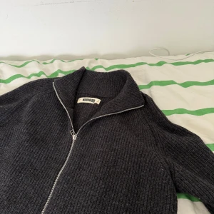 Mörkgrå stickad kofta från Woodbird - Säljer en mörkgrå stickad kofta från Woodbird med hel dragkedja och hög krage. Koftan är ribbstickad och har långa ärmar, perfekt för lager-på-lager under kyliga dagar. Enkel och stilren design som funkar till det mesta. Originalpris/Nyspris: 999 kr