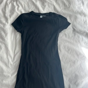 Svart kortklänning från H&M Divided - Enkel och stilren svart kortklänning från H&M Divided. Klänningen har rund halsringning, korta ärmar och är i en ribbad bomullsblandning som sitter snyggt och bekvämt. Perfekt basplagg att styla på många olika sätt.