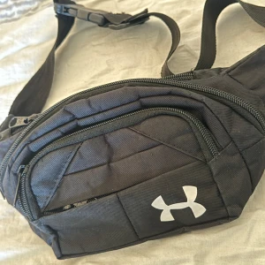 Svart magväska från Under Armour - Snygg svart magväska från Under Armour med justerbart band och två dragkedjefack. Väskan har en sportig look med vit logga framtill och är tillverkad i slitstark polyester. Perfekt för att bära det viktigaste när du är på språng.