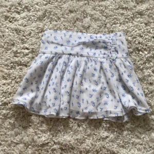 Vit blommig kortkjol från Gina Tricot - Supersöt vit kortkjol från Gina Tricot med ljusblått blommönster. Kjolen har strech i midjan och är lätt utställd för en flowy känsla. Perfekt för varma dagar och ger en fräsch sommarvibe.