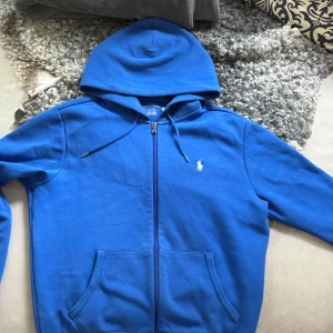 Blå hoodie från Polo Ralph Lauren - Snygg blå hoodie från Polo Ralph Lauren med dragkedja och klassisk huva. Tröjan har vit broderad logga på bröstet, känguruficka och justerbar snörning i huvan. Perfekt för chill dagar eller när du vill ha en clean streetstyle. Nypris runt 2000kr