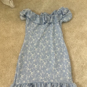 Blommig blå volangklänning Gina Tricot - Supersöt blå klänning från Gina Tricot med små vita blommor, volang vid axlarna och nertill. Offshoulder-modell som ger en somrig vibe. Perfekt för dig som gillar romantisk och trendig stil.