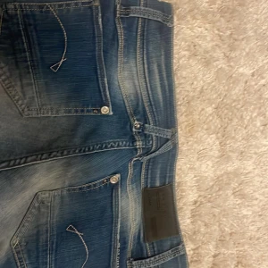 G-Star Raw jeansshorts blå denim - Snygga blå jeansshorts från G-Star Raw med råa, fransiga kanter och klassiska fem fickor. Shorts med slitningar och tydliga kontrastsömmar, samt G-Star-logga på fickan. Perfekta för en chill sommarlook.