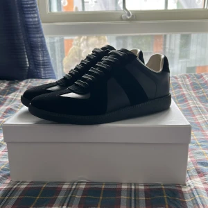 Maison Margiela gats - Maison Margiela gats sneakers i helsvart med både mocka och skinnpaneler. Klassisk siluett med rund tå, svarta snören och svart sula. Insidan är ljusbeige och skon har diskreta detaljer som gör dem riktigt stilrena och unika. 