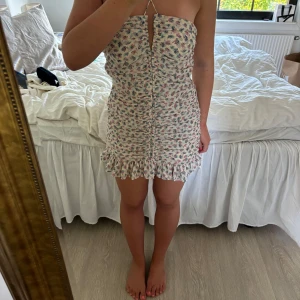 Blommig kort sommarklänning med volang - Supersöt kort klänning med blommigt mönster i rosa och blått. Axelbandslös modell med smala halterneckband, rynkade detaljer framtill och volangkant nertill. 