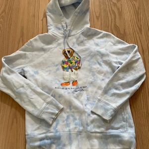 Polo Bear hoodie från Ralph Lauren M - Säljer en ljusblå och vit hoodie från Polo Ralph Lauren med ikoniska Polo Bear-trycket framtill. Tröjan har huva med snörning, känguruficka och mjukt bomullsmaterial. Perfekt för dig som gillar streetstyle och vill sticka ut med en klassisk look.