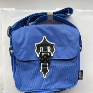 Blå axelväska från Trapstar - Säljer en blå axelväska från Trapstar med justerbart axelband och coolt svart-vitt emblem på locket. Väskan är gjord i slitstark polyester och har ett unikt spänne framtill. Perfekt för att bära dina essentials med stil.