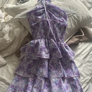 Lila blommig volangklänning med knyt - Supersöt kort klänning med lila och blått blommönster, volanger i flera lager och smockad överdel. Halterneck med knyt i nacken och lätt, luftig känsla. Perfekt för sommarens alla tillfällen!