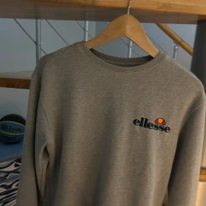 Ellese sweatshirt - Snygg grå sweatshirt från Ellesse med broderad logga på bröstet i blått, rött och gult. Klassisk rund halsringning och långa ärmar. Perfekt för en chill och sportig stil. Mjuk och skön bomullsblandning. Säljs då den inte används. Har till störst del bara hängd i garderoben. Nypris 800kr.