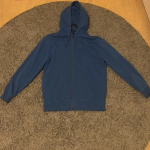Blå hoodie med dragkedja - En stilren blå hoodie med hel dragkedja framtill och två fickor. Tröjan har huva med snörning och långa ärmar. Perfekt för dig som gillar en enkel och clean look. Passar till både jeans och mjukisbyxor.