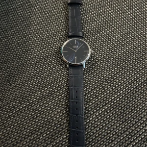 Svart klocka från GHW med läderband - Stilren klocka från GHW med svart urtavla och silverdetaljer. Klockan har ett klassiskt runt boett i silver och svart läderarmband med krokodilpräglad yta. Urtavlan är minimalistisk med datumvisning och tunna visare. Perfekt accessoar för en clean look. Den är helt ny, oanvänt den kostar i afären 999kr jag säljer för 450kr