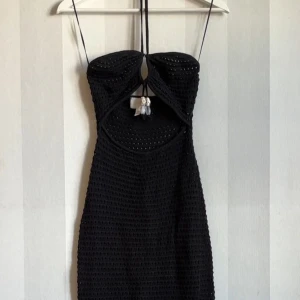 Svart virkad klänning från H&M - Svart virkad klänning  från H&M med snygg cut-out framtill och knyt i nacken. Klänningen är ärmlös och har pärldetaljer vid knytbandet. Perfekt för sommarens varma dagar och strandhäng.